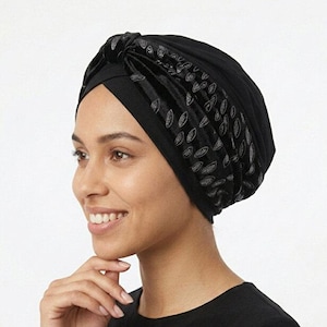 May include: Black turban with a velvet leaf pattern on the front. The turban is made of a soft material and is designed to be worn on the head. The person in the image is wearing a black long-sleeve shirt.