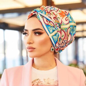 May include: A woman wearing a colorful headscarf with a floral and geometric design, a white t-shirt with the Guess logo, and a light pink blazer. The headscarf features shades of blue, red, and orange.