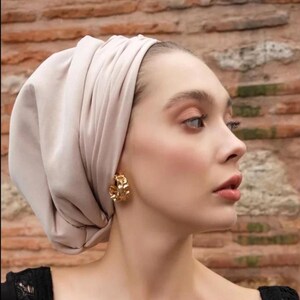 Può includere: Una donna con un velo beige e orecchini a cerchio dorati. Gli orecchini hanno un design floreale strutturato. Indossa un top nero con dettagli in pizzo. Lo sfondo è un muro di mattoni.