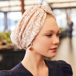 May include: A turban-style head covering in a cream color with a repeating geometric pattern in a light brown hue. The turban is worn with a dark blue top and a pair of amber-colored drop earrings.
