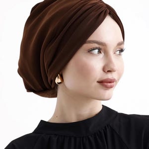 Puede incluir: Un turbante marr&oacute;n con un dise&ntilde;o fruncido. El cubrecabezas est&aacute; hecho de una tela suave y drapeada. La persona lleva pendientes dorados en forma de l&aacute;grima y una blusa negra.