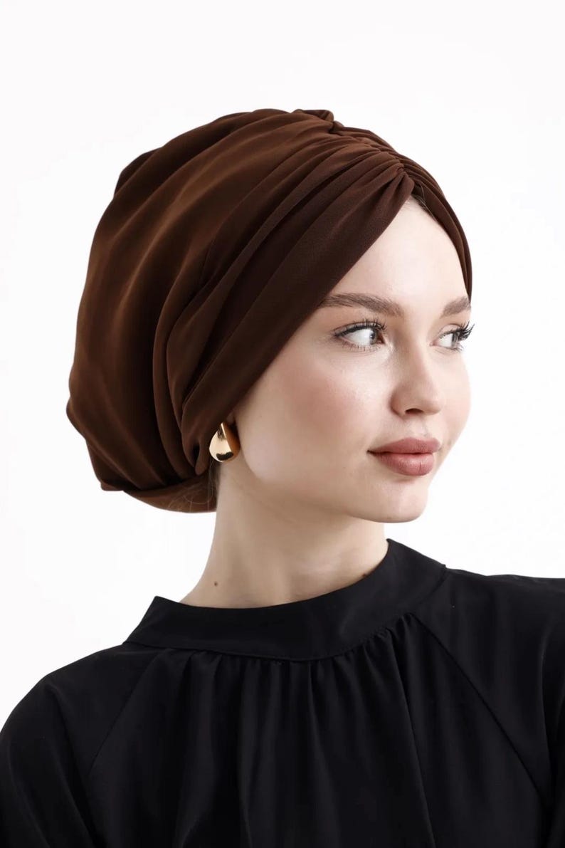 Puede incluir: Un turbante marr&oacute;n con un dise&ntilde;o fruncido. El cubrecabezas est&aacute; hecho de una tela suave y drapeada. La persona lleva pendientes dorados en forma de l&aacute;grima y una blusa negra.