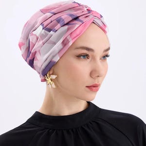 May include: A pink, purple, and white patterned turban. The head covering is worn by a person wearing gold flower earrings and a black top. The turban is a stylish accessory.