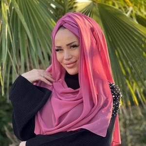 Può includere: Una donna indossa un hijab rosa e un maglione nero. L'hijab è drappeggiato sulla testa e sulle spalle, creando morbide pieghe. Il maglione ha un collo alto e maniche lunghe. Lo sfondo presenta foglie di palma verdi.