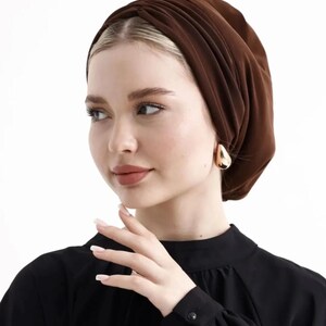 Puede incluir: Un turbante marr&oacute;n. La tela est&aacute; fruncida y drapeada, creando una apariencia texturizada. La persona lleva pendientes dorados en forma de l&aacute;grima y una blusa negra de manga larga. El fondo es blanco liso.