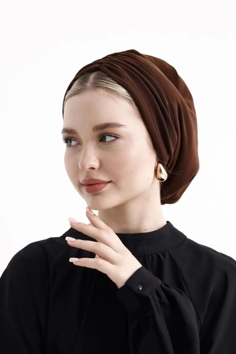 Puede incluir: Un turbante marr&oacute;n. La tela est&aacute; fruncida y drapeada, creando una apariencia texturizada. La persona lleva pendientes dorados en forma de l&aacute;grima y una blusa negra de manga larga. El fondo es blanco liso.