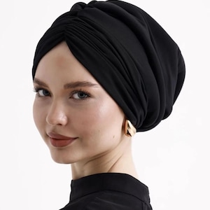 May include: A black turban-style head covering. The fabric is draped and gathered, creating a textured appearance. The person is wearing gold-coloured earrings and a black top. The background is plain white.
