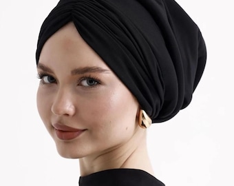 Puede incluir: Un turbante negro estilo turbante. La tela está drapeada y fruncida, creando una apariencia texturizada. La persona lleva pendientes dorados y una blusa negra. El fondo es blanco liso.