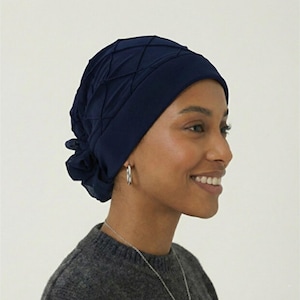 May include: A navy blue head covering with a quilted design. The head covering is made of a soft fabric and has a gathered detail on the side. The person in the image is wearing a gray sweater and silver hoop earrings.