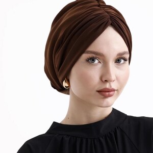 Puede incluir: Un turbante marr&oacute;n con un dise&ntilde;o fruncido. El tocado est&aacute; hecho de una tela suave y se usa con una blusa negra y aretes dorados. El turbante es un accesorio elegante.