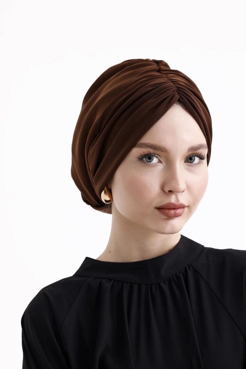 Puede incluir: Un turbante marr&oacute;n con un dise&ntilde;o fruncido. El tocado est&aacute; hecho de una tela suave y se usa con una blusa negra y aretes dorados. El turbante es un accesorio elegante.