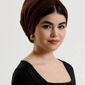 Puede incluir: Una mujer con un turbante marr&oacute;n oscuro y pendientes de aro dorados. Lleva una camiseta negra con cuello cuadrado. El turbante est&aacute; hecho de una tela suave y drapeada.