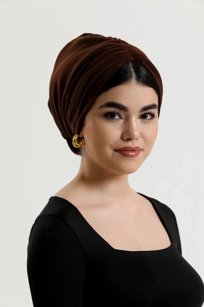 Puede incluir: Una mujer con un turbante marr&oacute;n oscuro y pendientes de aro dorados. Lleva una camiseta negra con cuello cuadrado. El turbante est&aacute; hecho de una tela suave y drapeada.