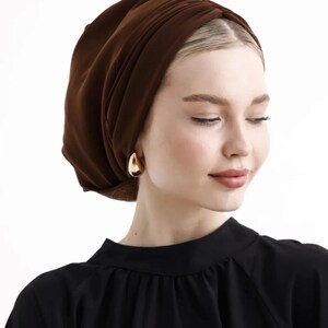 Puede incluir: Una mujer con un turbante marr&oacute;n y pendientes dorados en forma de l&aacute;grima. Lleva una blusa negra. El turbante es de tela suave y drapeada. El fondo es blanco liso.