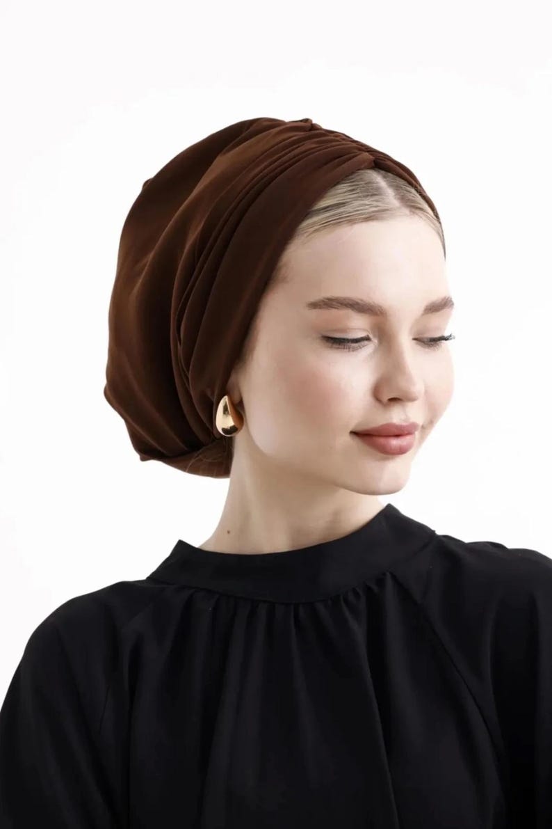 Puede incluir: Una mujer con un turbante marr&oacute;n y pendientes dorados en forma de l&aacute;grima. Lleva una blusa negra. El turbante es de tela suave y drapeada. El fondo es blanco liso.