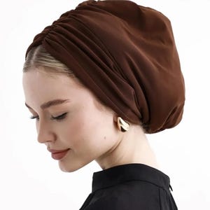 Puede incluir: Un turbante marr&oacute;n con detalles de tela fruncida. El tocado lo lleva una persona que lleva pendientes dorados y una parte superior negra. El fondo es blanco.