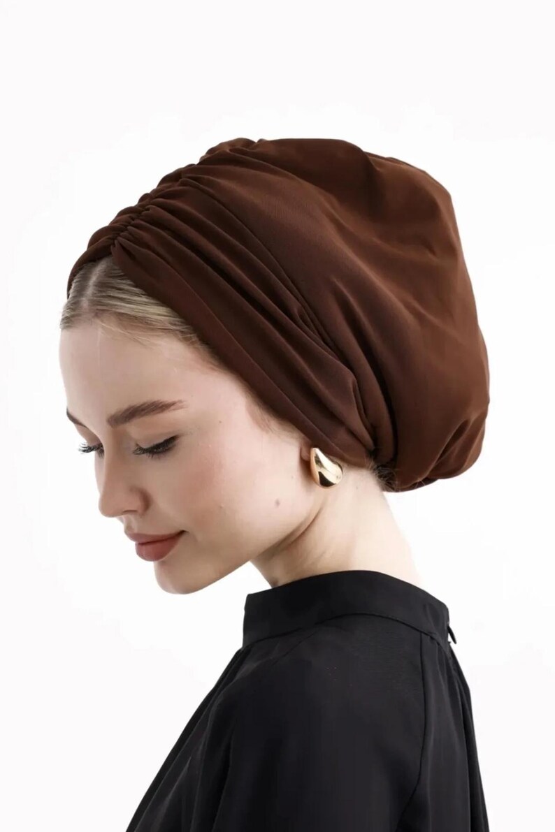Puede incluir: Un turbante marr&oacute;n con detalles de tela fruncida. El tocado lo lleva una persona que lleva pendientes dorados y una parte superior negra. El fondo es blanco.