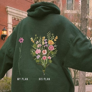 Könnte beinhalten: Ein dunkelgrüner Kapuzenpullover mit floraler Stickerei auf dem Rücken. Die Stickerei zeigt einen Strauß bunter Blumen und den Text "MY PLAN" und "HIS PLAN". Die Person trägt eine hellblaue Jeans und weiße Turnschuhe.