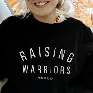 May include: A black sweatshirt with the text "RAISING WARRIORS" and "PSALM 127:4" in white. The person is wearing a grey beanie, black sunglasses, and a gold chain necklace. They are sitting in a car and wearing plaid trousers.