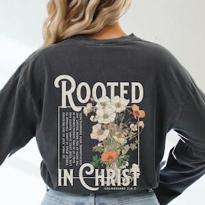 以下が含まれることがあります： 花柄のデザインと「Rooted in Christ Colossians 2:6-7」というテキストが書かれた、長袖の濃いグレーのTシャツです。