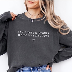 May include: A dark gray long-sleeved shirt with the text "Can't throw stones while washing feet" and a small cross symbol.