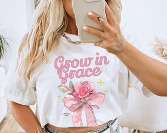 May include: A white t-shirt with a pink graphic that says "Grow in Grace" with a pink rose and bow. The person is wearing a light blue denim skirt and a white belt.