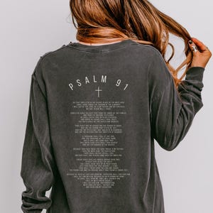 May include: Dark grey long-sleeved shirt with the text "PSALM 91" and a cross graphic on the back. The shirt has a distressed, vintage appearance. The text is in a simple, sans-serif font. A casual wear item.