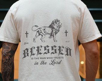 May include: Cream-colored t-shirt with a lion graphic and the text "BLESSED is the man who trusts in the Lord." The design includes crosses and the text "JER 17:7". The shirt is a casual wear item.