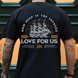 May include: Black t-shirt with a graphic design on the back. The design features a sailing ship, the text "HOW DEEP IS THE FATHERS LOVE FOR US", and the Bible verse Ephesians 3:18-19. The shirt is worn by a person.