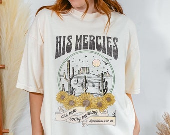 May include: A cream colored t-shirt with a graphic of a desert landscape with cacti and sunflowers. The text "His Mercies are new every morning" is printed on the shirt with a scripture reference.
