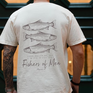 Puede incluir: Camiseta color crema con tres ilustraciones de peces apiladas verticalmente. Debajo de los peces, el texto dice: "COME, FOLLOW ME, AND I WILL MAKE YOU Fishers of Men Mark III."