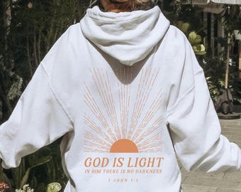 May include: White hoodie with an orange sunburst design and the text "GOD IS LIGHT IN HIM THERE IS NO DARKNESS 1 JOHN 1:5". The hoodie is worn by a person standing outside near plants.