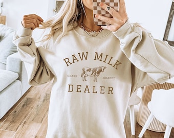 May include: A beige sweatshirt with the text "RAW MILK GRASS GRAZED DEALER" and a brown cow illustration. The model is wearing the sweatshirt with brown shorts and white sneakers.