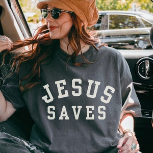 May include: A woman wearing a gray t-shirt with the text "JESUS SAVES" in white lettering. She is sitting in a car and wearing sunglasses and a brown hat.