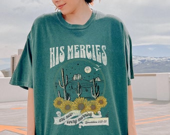 May include: A green t-shirt with a graphic print of a desert scene with cacti and sunflowers. The text "His Mercies are new every morning" is printed on the shirt.