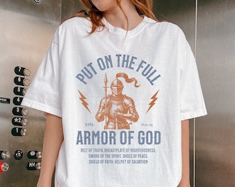 Op de afbeelding: Wit T-shirt met een afbeelding van een ridder in harnas en de tekst "Put on the Full Armor of God" met bijbelverzen. De tekst onder de afbeelding luidt "Gordel van de waarheid, borstplaat van gerechtigheid, zwaard van de Geest, schoenen van vrede, schild van het geloof, helm van de zaligheid."