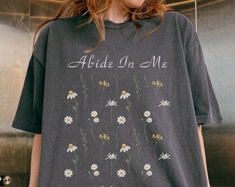 Puede incluir: Una camiseta negra de gran tamaño con un estampado de margaritas blancas y el texto "Abide In Me" y "John 15:4".