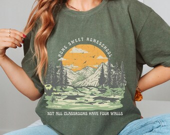 May include: Olive green t-shirt with a landscape graphic featuring mountains, trees, and a sunset. Text reads "HOME SWEET HOMESCHOOL" above the graphic and "NOT ALL CLASSROOMS HAVE FOUR WALLS" below.