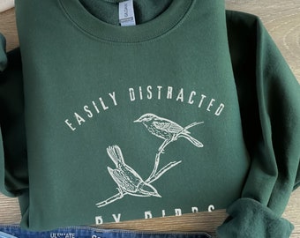 May include: A dark green sweatshirt with the text "Easily Distracted by Birds" in white lettering. Two birds are illustrated on a branch above the text.