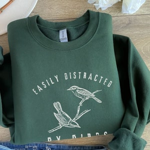 May include: A dark green sweatshirt with the text "Easily Distracted by Birds" in white lettering. Two birds are illustrated on a branch above the text.