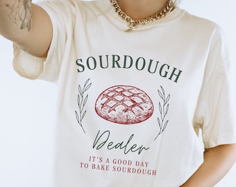 May include: A white t-shirt with the text "SOURDOUGH Dealer" and a drawing of a loaf of bread. The text below the bread says "It's a good day to bake sourdough."