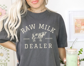 May include: A dark gray t-shirt with a vintage-style graphic that reads "RAW MILK DEALER" with a stylized cow illustration and the words "GRASS GRAZED" underneath.