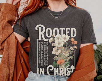 Könnte beinhalten: Ein schwarzes T-Shirt mit einem Vintage-Grafikdesign, das einen Blumenstrauß und den Text "Rooted in Christ" zeigt. Der Text ist in weißer Schrift mit schwarzem Umriss. Der Blumenstrauß ist in Weiß-, Orange- und Grüntönen gehalten.