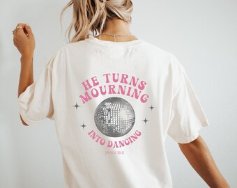 May include: White t-shirt with a pink and gray graphic that says "He turns mourning into dancing" with a disco ball and the text "Psalm 30:11".
