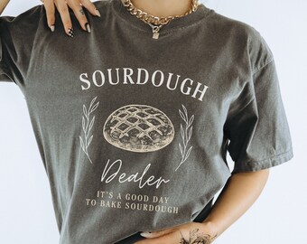 May include: A gray t-shirt with the text "SOURDOUGH Dealer" and a drawing of a loaf of bread. The text below the bread says "It's a good day to bake sourdough."