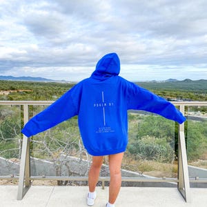 May include: A vibrant blue hoodie with the text "PSALM 81" and a cross design on the back. The hoodie also features additional text below the cross. The person is wearing white sneakers and socks.