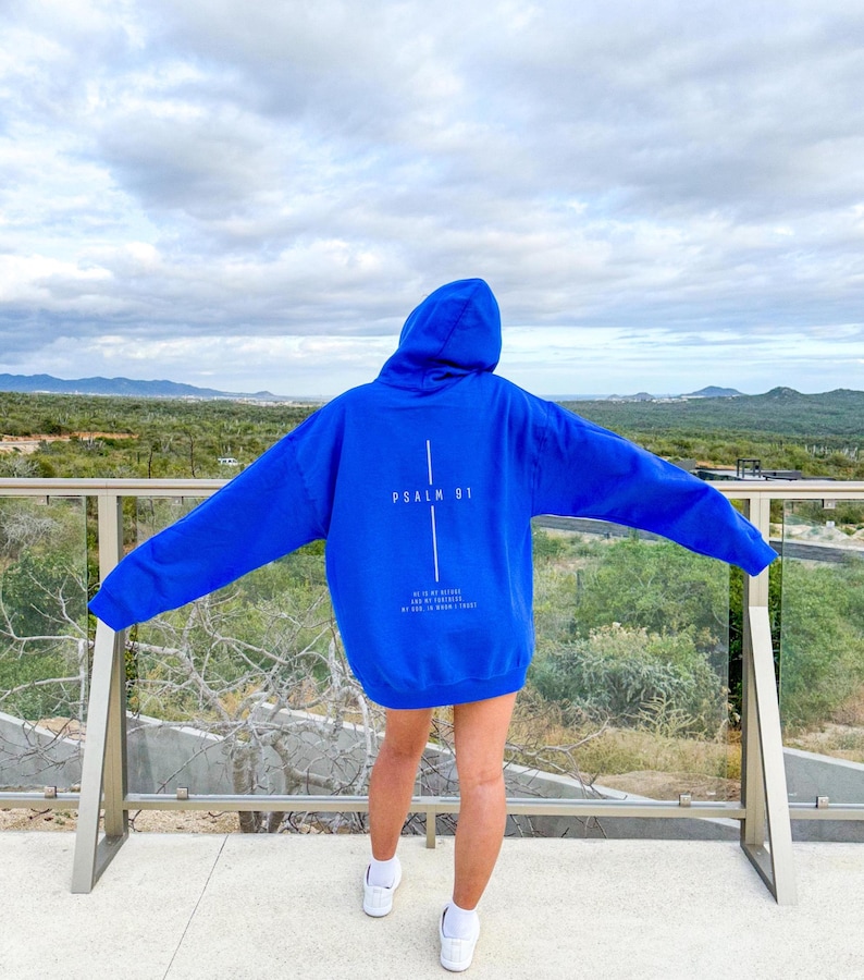 May include: A vibrant blue hoodie with the text "PSALM 81" and a cross design on the back. The hoodie also features additional text below the cross. The person is wearing white sneakers and socks.