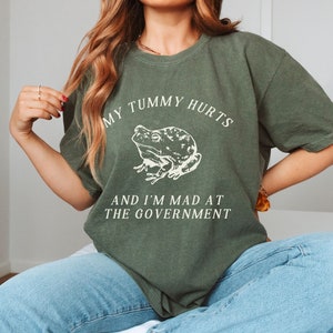 May include: A green t-shirt with a white frog graphic and the text "My tummy hurts and I'm mad at the government."