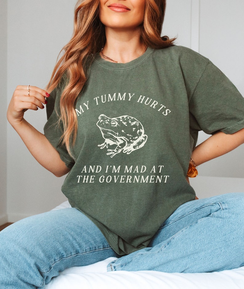May include: A green t-shirt with a white frog graphic and the text "My tummy hurts and I'm mad at the government."