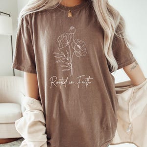 Puede incluir: Una camiseta marr&oacute;n de gran tama&ntilde;o con un contorno blanco de flores y el texto "Rooted in Faith".